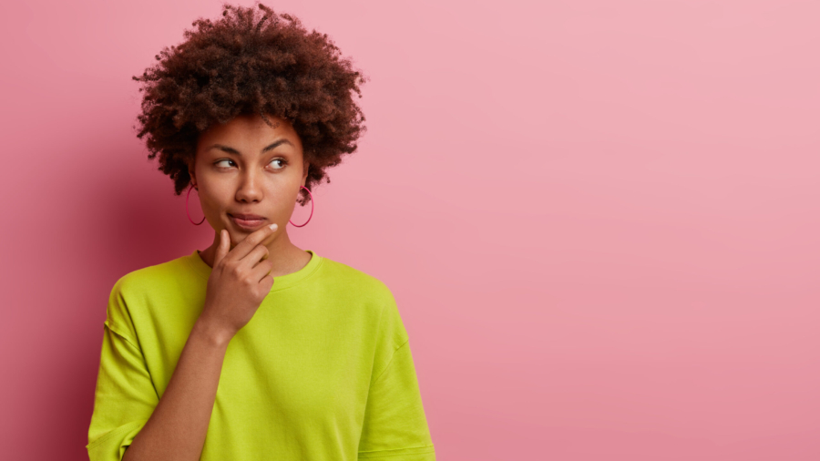 Horizontal shot of pleasant looking Afro American woman holds chin and looks pensively away, thinks about great plan, has new ideas in mind, wears bright green t shirt, poses over rosy wall copy space